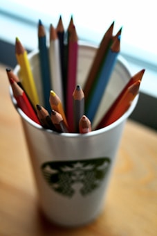 A collection of colorful wooden pencils is placed inside a paper cup. The cup features a distinct logo and sits on a wooden surface. The pencils vary in color, including red, yellow, blue, and orange, and are arranged with their sharpened tips pointing upward. A soft, diffused light brightens the scene, adding warmth to the overall atmosphere.