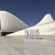 A modern architectural structure with smooth, flowing lines and a sleek white exterior stands against a clear blue sky. A large sign in front features bold, geometric text.