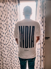 A person is standing with their back to the camera, wearing a white t-shirt that has the word 'INDIA' in large bold letters and 'Outcast' below it. The person is standing in front of a window with sheer, patterned curtains that have a dark blue dot design.