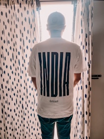 A person is standing with their back to the camera, wearing a white t-shirt that has the word 'INDIA' in large bold letters and 'Outcast' below it. The person is standing in front of a window with sheer, patterned curtains that have a dark blue dot design.