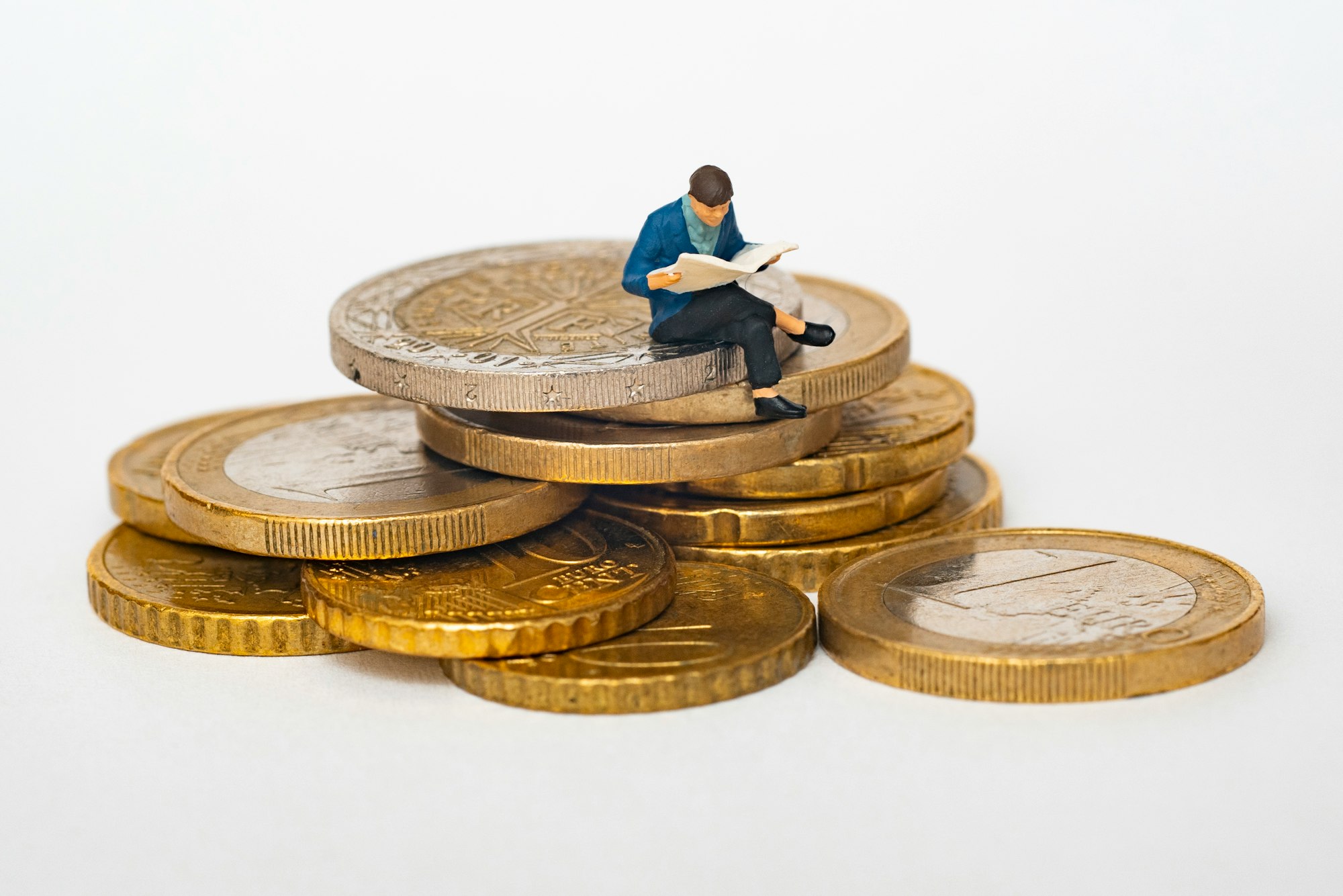 personnage avec une veste bleue lisant le journal assis sur une pile de pièces de monnaie euro sur un fond blanc.