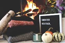 A cozy arrangement featuring a stack of folded sweaters in various colors next to a small pile of birch logs in front of a crackling fire. A black letter board with the message 'Sweater Weather' is placed nearby, alongside a green candle and a trio of small pumpkins, with a vibrant bouquet of flowers.