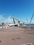 A large solar panel structure is positioned prominently under a clear blue sky, with people walking nearby. The foreground features a plaza area with patterned paving and colorful barriers, while in the background, a marina with several masts is visible.