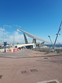 A large solar panel structure is positioned prominently under a clear blue sky, with people walking nearby. The foreground features a plaza area with patterned paving and colorful barriers, while in the background, a marina with several masts is visible.