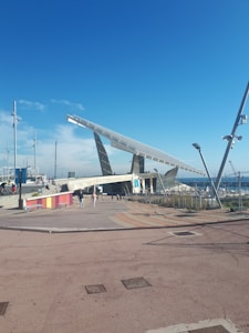 A large solar panel structure is positioned prominently under a clear blue sky, with people walking nearby. The foreground features a plaza area with patterned paving and colorful barriers, while in the background, a marina with several masts is visible.