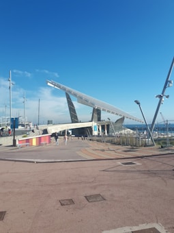 A large solar panel structure is positioned prominently under a clear blue sky, with people walking nearby. The foreground features a plaza area with patterned paving and colorful barriers, while in the background, a marina with several masts is visible.