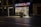 A dimly lit street scene featuring a small shop with brightly illuminated windows displaying various goods. Neon signs above the store advertise 'FRUTOS SECOS' and 'Nuts and Snacks'. A trash bin is placed on the sidewalk near the storefront. Nearby, there are closed neighboring shops with darkened windows.
