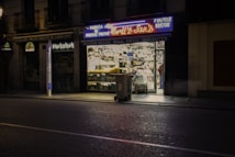 A dimly lit street scene featuring a small shop with brightly illuminated windows displaying various goods. Neon signs above the store advertise 'FRUTOS SECOS' and 'Nuts and Snacks'. A trash bin is placed on the sidewalk near the storefront. Nearby, there are closed neighboring shops with darkened windows.