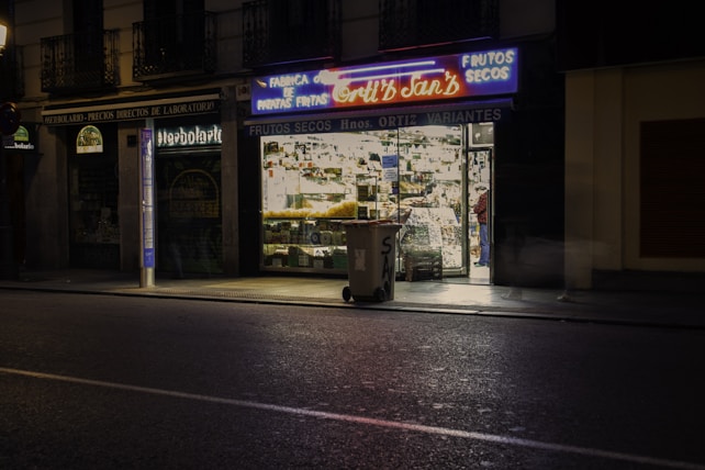 A dimly lit street scene featuring a small shop with brightly illuminated windows displaying various goods. Neon signs above the store advertise 'FRUTOS SECOS' and 'Nuts and Snacks'. A trash bin is placed on the sidewalk near the storefront. Nearby, there are closed neighboring shops with darkened windows.