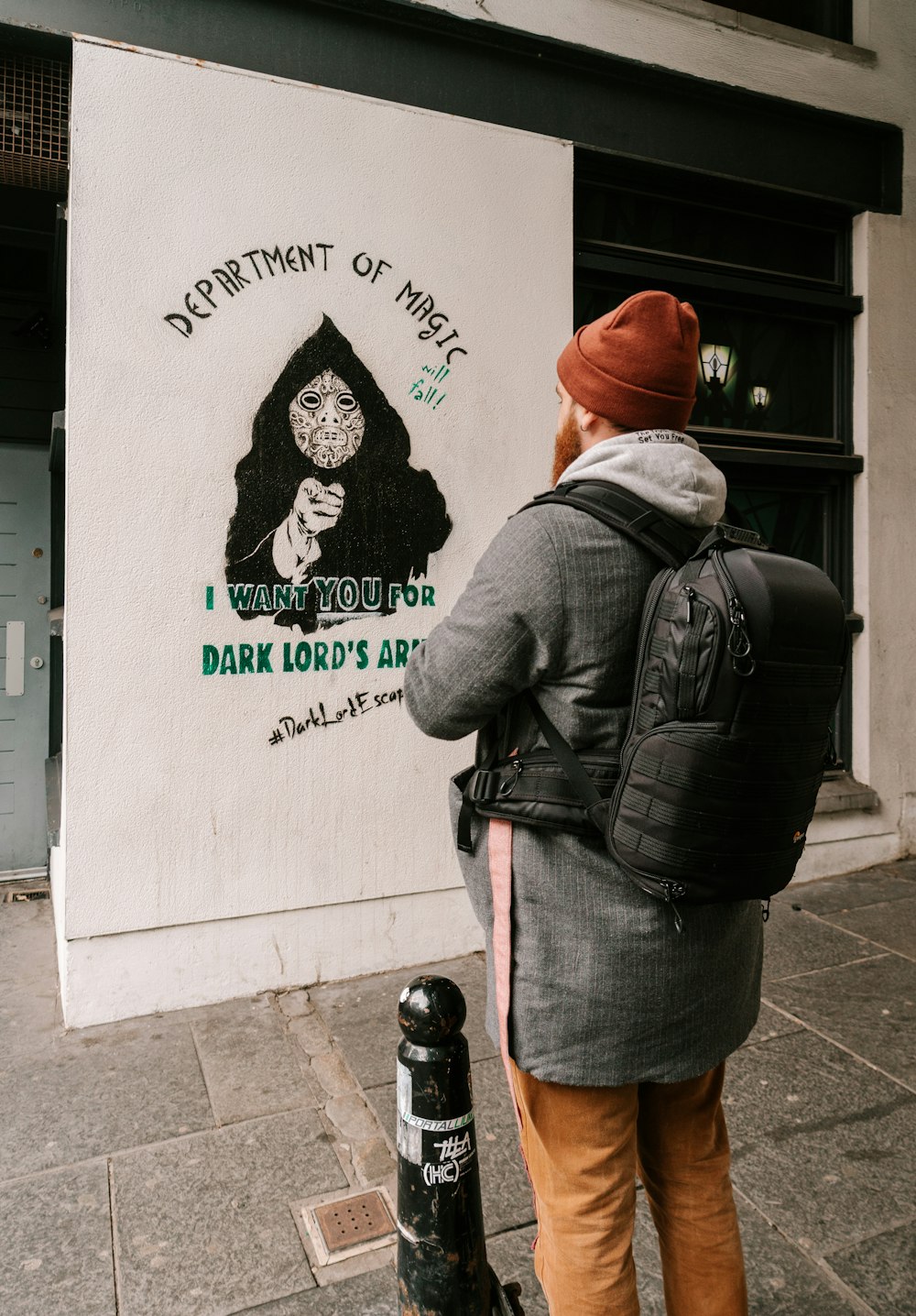 man in black leather jacket and brown hat standing beside white wall