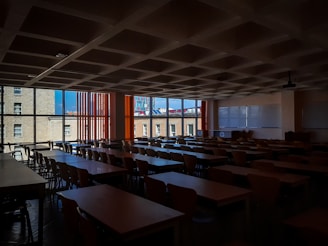 A dimly lit classroom filled with rows of empty wooden desks and chairs. Large windows on the left side let in natural light, revealing buildings outside. There are two large whiteboards on the wall to the right, and a ceiling-mounted projector is also visible.