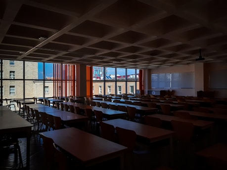 A dimly lit classroom filled with rows of empty wooden desks and chairs. Large windows on the left side let in natural light, revealing buildings outside. There are two large whiteboards on the wall to the right, and a ceiling-mounted projector is also visible.