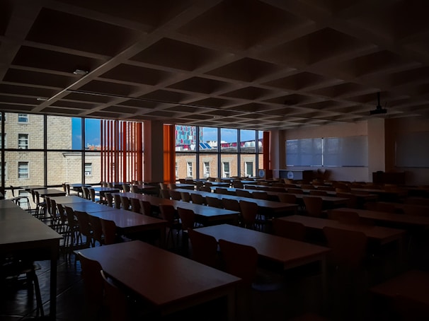 A dimly lit classroom filled with rows of empty wooden desks and chairs. Large windows on the left side let in natural light, revealing buildings outside. There are two large whiteboards on the wall to the right, and a ceiling-mounted projector is also visible.