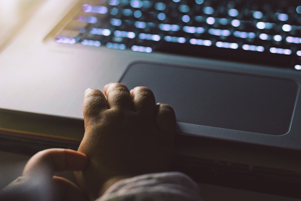 A small child's hand gently rests on the trackpad of a laptop. The surrounding area is dimly lit, with the laptop keyboard illuminated by a soft glow from its backlight. The scene suggests a quiet, intimate atmosphere, possibly of a child exploring technology.