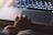 A small child's hand gently rests on the trackpad of a laptop. The surrounding area is dimly lit, with the laptop keyboard illuminated by a soft glow from its backlight. The scene suggests a quiet, intimate atmosphere, possibly of a child exploring technology.