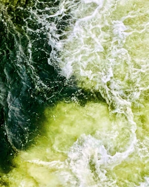 An aerial view of turbulent water, with swirling currents and frothy white waves creating dynamic patterns. The water appears to be moving rapidly, with a contrast between darker and lighter areas.