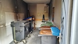 A small, dimly lit storage area or room containing several large trash bins, some with lids in shades of brown and green. The walls and floor show signs of wear and grime, and there are cleaning tools, such as a broom and mop, stored against one wall. A ventilation fan and a window are visible at the back.