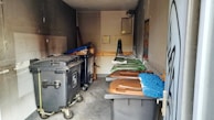 A small, dimly lit storage area or room containing several large trash bins, some with lids in shades of brown and green. The walls and floor show signs of wear and grime, and there are cleaning tools, such as a broom and mop, stored against one wall. A ventilation fan and a window are visible at the back.