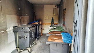 A small, dimly lit storage area or room containing several large trash bins, some with lids in shades of brown and green. The walls and floor show signs of wear and grime, and there are cleaning tools, such as a broom and mop, stored against one wall. A ventilation fan and a window are visible at the back.