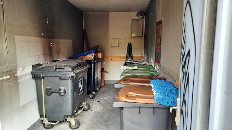 A small, dimly lit storage area or room containing several large trash bins, some with lids in shades of brown and green. The walls and floor show signs of wear and grime, and there are cleaning tools, such as a broom and mop, stored against one wall. A ventilation fan and a window are visible at the back.