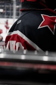 A close-up of a hockey jersey, primarily featuring the shoulder and back area with a red and white star design. The name 'Lutch' is visible on the shoulder. The background shows a blurry scene with indistinct figures, likely other players, on an ice rink.