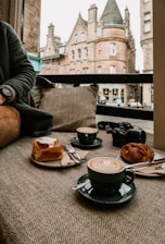 A cozy café setting with two cappuccinos adorned with latte art, alongside a cinnamon roll and a pastry. A camera is placed next to the pastries, and in the background, large historic buildings with intricate architecture are visible through a window.