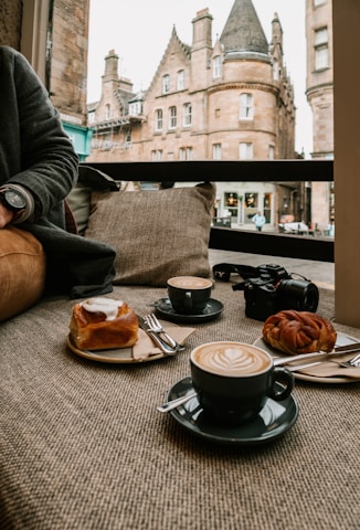A cozy café setting with two cappuccinos adorned with latte art, alongside a cinnamon roll and a pastry. A camera is placed next to the pastries, and in the background, large historic buildings with intricate architecture are visible through a window.