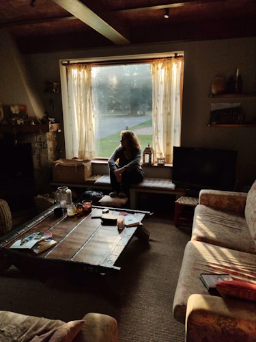A cozy living room is softly illuminated by the light filtering through the window. A person is sitting on a bench by the window, surrounded by warm tones and rustic decor. The wooden coffee table holds various items like magazines, candles, and a glass jar. The seating area includes a patterned couch and a view of the outdoors through semi-transparent curtains.