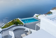 Overlooking a serene infinity pool, the scene features crisp white architecture cascading down towards the calm blue sea. The area is adorned with green plants and a small boat is visible on the water. The setting exudes luxury and tranquility.