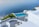 Overlooking a serene infinity pool, the scene features crisp white architecture cascading down towards the calm blue sea. The area is adorned with green plants and a small boat is visible on the water. The setting exudes luxury and tranquility.