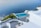 Overlooking a serene infinity pool, the scene features crisp white architecture cascading down towards the calm blue sea. The area is adorned with green plants and a small boat is visible on the water. The setting exudes luxury and tranquility.