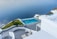 Overlooking a serene infinity pool, the scene features crisp white architecture cascading down towards the calm blue sea. The area is adorned with green plants and a small boat is visible on the water. The setting exudes luxury and tranquility.
