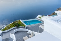 Overlooking a serene infinity pool, the scene features crisp white architecture cascading down towards the calm blue sea. The area is adorned with green plants and a small boat is visible on the water. The setting exudes luxury and tranquility.