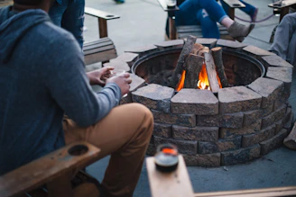 A group of people are sitting around a stone fire pit with a fire burning in the center. The scene suggests a casual and relaxed atmosphere, as people appear to be enjoying a cozy outdoor gathering.