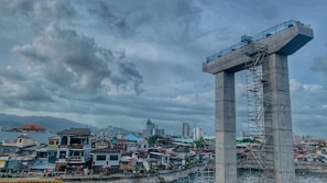 A construction site with a large concrete structure resembling a tall unfinished bridge pier is visible. Scaffolding runs along its side. In the background, a densely packed urban area with a variety of residential buildings and a few taller skyscrapers. The sky is overcast with thick clouds.