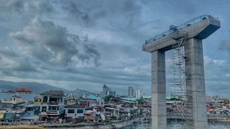 A construction site with a large concrete structure resembling a tall unfinished bridge pier is visible. Scaffolding runs along its side. In the background, a densely packed urban area with a variety of residential buildings and a few taller skyscrapers. The sky is overcast with thick clouds.