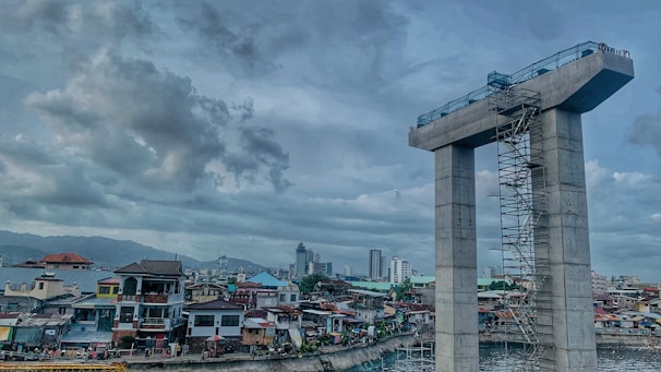 A construction site with a large concrete structure resembling a tall unfinished bridge pier is visible. Scaffolding runs along its side. In the background, a densely packed urban area with a variety of residential buildings and a few taller skyscrapers. The sky is overcast with thick clouds.
