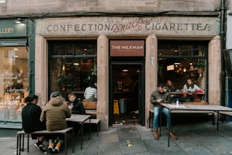 A cozy café with an old-fashioned sign reading 'Confections, Snacks, Cigarettes' above the entrance. People are sitting at wooden benches outside, some engaged in conversation and others on their phones. Through the window, more patrons can be seen inside enjoying their time. The atmosphere looks relaxed and inviting.