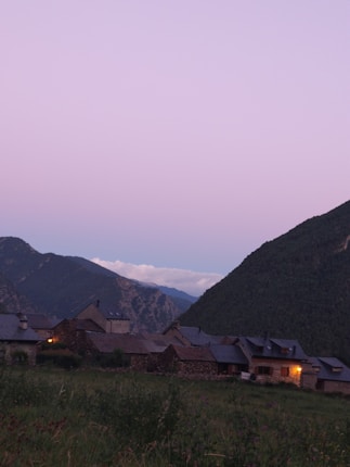 A serene village nestled in the mountains, showcasing rustic stone houses with warm lights glowing from the windows. The surrounding landscape includes lush green hills and a sky with a soft pink hue, suggesting early evening or dawn.