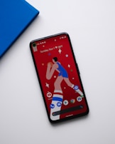 A smartphone with a colorful screen displaying a cartoon image of a basketball player and several app icons at the bottom. The phone is placed on a white surface next to a blue book or notebook.