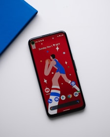 A smartphone with a colorful screen displaying a cartoon image of a basketball player and several app icons at the bottom. The phone is placed on a white surface next to a blue book or notebook.