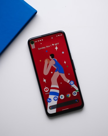 A smartphone with a colorful screen displaying a cartoon image of a basketball player and several app icons at the bottom. The phone is placed on a white surface next to a blue book or notebook.