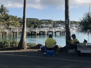 Several people wearing yellow high-visibility jackets are sitting in a shaded area near a waterfront. In the background, there is a marina with numerous docked boats and yachts. Palm trees and other greenery provide a tropical feel. The scene is relaxed, with a clear sky and calm water.