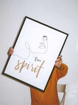 A person in an orange sweater holds a framed artwork featuring a minimalist line drawing of a face and the phrase 'free spirit' written in golden glittery letters.