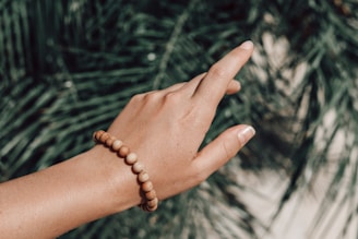 A hand wearing a wooden beaded bracelet is extended against a background of lush green foliage. The hand has a natural skin tone and well-kept nails, adding a subtle elegance to the scene.