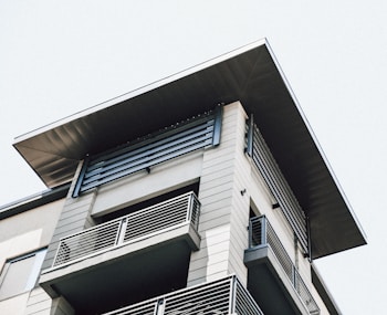 A modern building with a flat roof and multiple balconies featuring metal railings. The architecture includes clean lines and a combination of light and dark materials, creating a contemporary design.