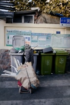 A street scene with several recycling bins, each designated for a specific type of waste, against a wall adorned with informational posters about recycling. In front of the bins is a cart filled with assorted materials such as cardboard, plastic, and other recyclables. Above, a stone wall and foliage are visible, and there are street signs with directional information.