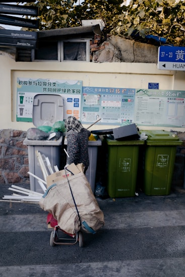 A street scene with several recycling bins, each designated for a specific type of waste, against a wall adorned with informational posters about recycling. In front of the bins is a cart filled with assorted materials such as cardboard, plastic, and other recyclables. Above, a stone wall and foliage are visible, and there are street signs with directional information.