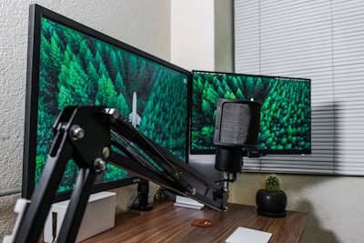 A workspace featuring two computer monitors displaying lush green forest images. A microphone with an adjustable arm sits prominently in front of the monitors. The desk is wooden with a small potted plant and a closed window blind in the background.