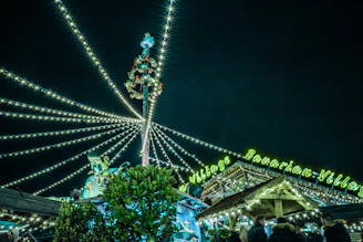 A festive night scene featuring a Bavarian-themed village decorated with an abundance of string lights. A tall pole with a wreath and decorations is centrally located, with lights radiating outward. Neon green signage displays 'Bavarian Village' above wooden structures and people in warm clothing.